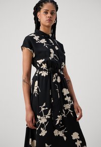 Melnas kleitas ar īsām piedurknēm, apkakles dekoltē, krēma ziedu raksts, josta vidukļa piekļaušanai, gluda auduma tekstūra, plūstoša silueta.