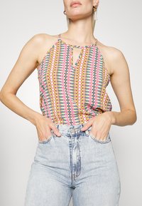 Top sem mangas colorido com um padrão geométrico em rosa, amarelo e azul. Decote em forma de lágrima, tecido leve. Combinado com jeans de cintura alta.