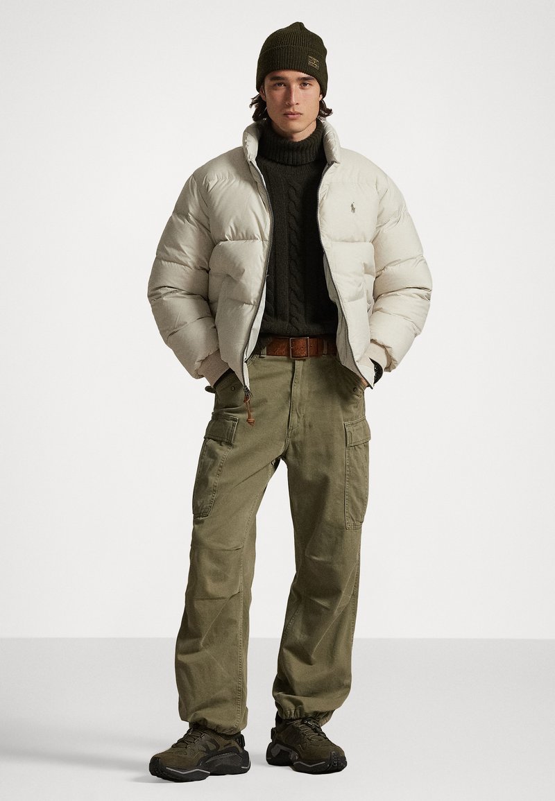 Dik crèmekleurige jas over een donkergroene trui, olijfgroene cargobroek en zwarte sneakers. Heeft een gebreide beanie en bruine riemaccenten.