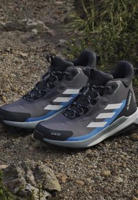 Ζευγάρι γκρι και μπλε παπούτσια πεζοπορίας Adidas Terrex σε βραχώδες εξωτερικό έδαφος με κοντινά πράσινα φυτά.
