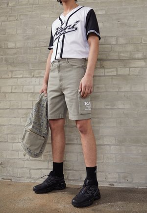 Hvid og sort baseball-jersey med "Voni" tekst, grå cargoshorts med sidelommer, sorte sko og mønstret taske.