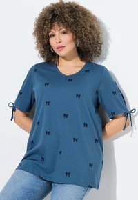 Haut bleu à manches courtes en tissu doux, avec un décolleté en V et des motifs de nœuds noirs décoratifs. Des liens sur les manches ajoutent des détails.