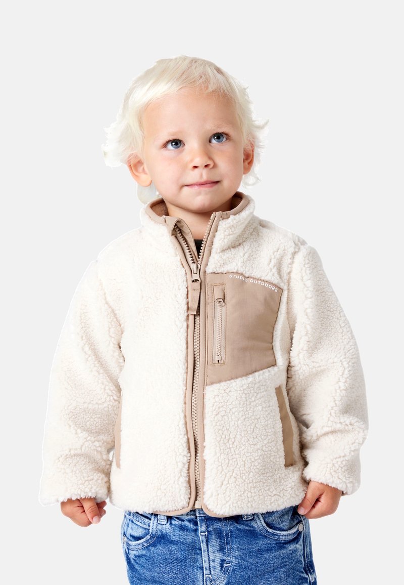 Tout-petit blond portant une veste en polaire blanche avec des poches beige et un jean bleu, debout et regardant légèrement vers le haut.