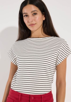 Junge Frau mit langen dunklen Haaren, die ein weißes Shirt mit schwarzen horizontalen Streifen und rote Hose trägt, schaut mit neutralem Gesichtsausdruck nach vorne.