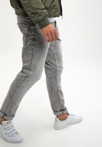 Jeans de mezclilla gris con corte ajustado, que presentan un aspecto desgastado y puños enrollados, combinados con zapatillas blancas y una chaqueta bomber de color verde olivo.