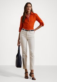 Orange linen shirt, white fitted jeans, navy belt with logo, black handbag, and black strappy high-heeled sandals. Model stands against a plain background.