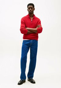 Pull en maille rouge à fermeture éclair avec des poignets côtelés, associé à un jean droit bleu et des chaussures en cuir noir, mannequin se tenant debout les bras croisés.