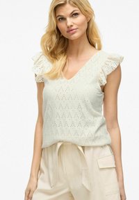 Hvid ærmeløs top med flæser på skuldrene, der har et struktureret stof med diamantudskæringer. V-halsdesign, bæres med lysebeige bukser.