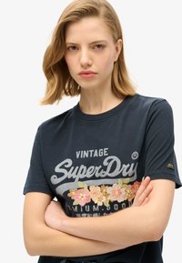 T-shirt in cotone blu navy con maniche corte, caratterizzata da un logo vintage SuperDry e ricami floreali in colori pastello sul petto.