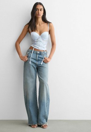 Kvinde iført en lyseblå blonder-bustier og højtaljede vidde-blå jeans, stående med hænderne nær lommerne mod en ensfarvet hvid væg.