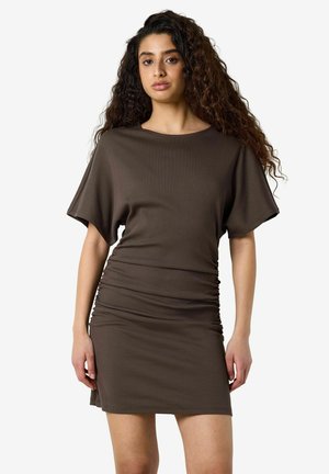 Woman with long curly hair wearing a short brown dress with ruched sides, standing against a plain light background.