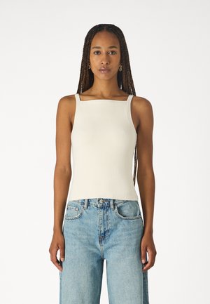 Ribbed, cream-colored top tankki, jossa on neliön muotoinen kaula-aukko ja olkaimet; yhdistetty korkeavyötäröisiin vaaleansinisiin farkkuihin rennon istuvuuden saamiseksi.