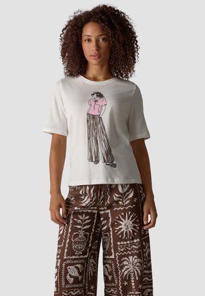 Woman with curly hair wearing a white t-shirt with a graphic of a woman and brown wide-leg pants with white nature-themed patterns.