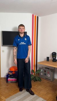 Μπλε ποδοσφαιρική φανέλα με το κείμενο «TRUE COLOURS», μαύρες λεπτομέρειες και κλασικό γιακά, συνδυασμένη με φαρδιά σκούρα τζιν παντελόνια και καφέ παπούτσια.