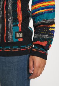 Musta neulepaita, jossa on monivärisiä kuvioita, teksturoitu muotoilu, etiketti vasemmassa alareunassa ja resorihihansuut. Yhdistetty sinisiin farkkuihin.