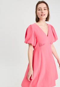 Robe rose avec un décolleté en V et des manches bouffantes, fabriquée en tissu doux avec une jupe évasée, texture lisse et sans motifs visibles.