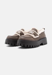 Bruine leren schoenen met witte nepbont voering, robuuste zwarte rubberen zool en metalen gesp detail met de merknaam.