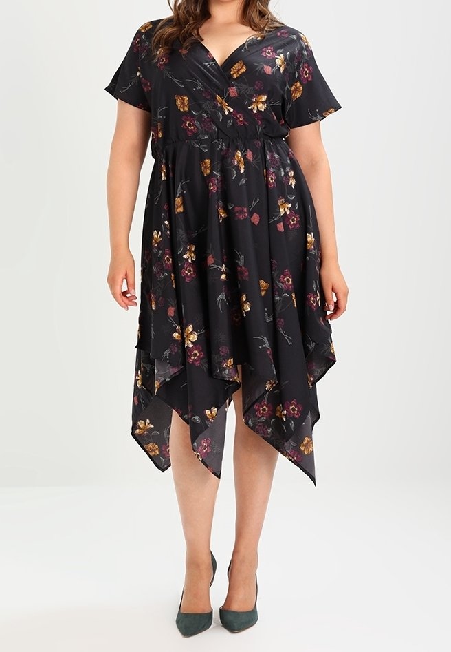 Femme portant une robe noire à motifs floraux avec manches courtes et un ourlet asymétrique en forme de mouchoir, associée à des chaussures pointues vert foncé.