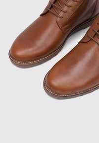 Braune Lederstiefel mit glatter Textur, runder Spitze und Schnürdesign. Verfügt über kontrastierender weißer Naht entlang der Sohle.
