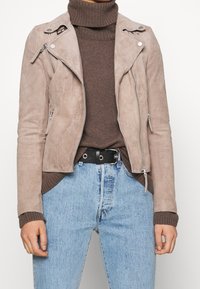 Bunda na motorku z taupe semiše s asymetrickým zipem, perforovanou texturou a stříbrnými kovovými prvky, spojená s hnědým rolákem a džíny z denimu.