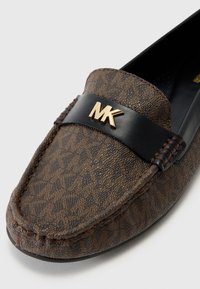 Bruine loafers met een gestructureerde stoffen bovenkant, een zwarte leren band met een gouden MK-logo en gestikte details langs de randen.