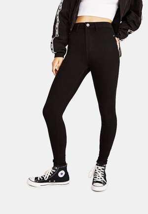 Femme portant un jean skinny taille haute noir, des baskets montantes noires et blanches, un top blanc court, et une veste zippée noire avec des bandes sur les manches.