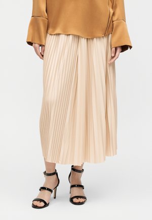 Γυναίκα που φοράει μπεζ πλισέ μίντι φούστα, μουσταρδί μπλούζα με μακριά μανίκια και μαύρα πέδιλα με ψηλό τακούνι, ανοιχτά στα δάχτυλα και με λουριά στον αστράγαλο.