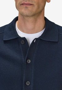 Marinblå ribbad stickad cardigan med krage, knappstängning och strukturerad design. Lager på en vit skjorta.