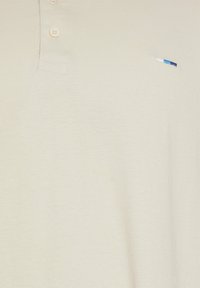 Polo beige avec deux boutons et un petit logo brodé bleu, blanc et bleu foncé sur la poitrine.