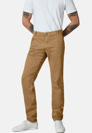 Mann trägt beige Freizeithose, weißes T-Shirt, weiße Sneakers, steht mit den Händen hinter dem Rücken, sichtbare Arm-Tattoos.