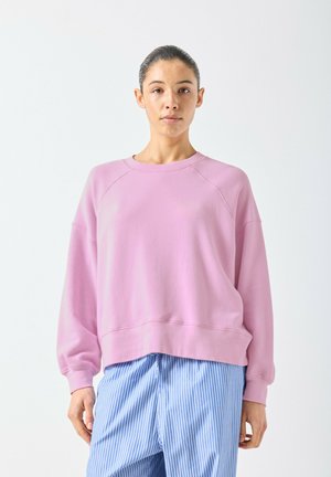 Donna che indossa una felpa rosa larga e pantaloni a righe blu e bianche, in piedi su uno sfondo bianco uniforme.