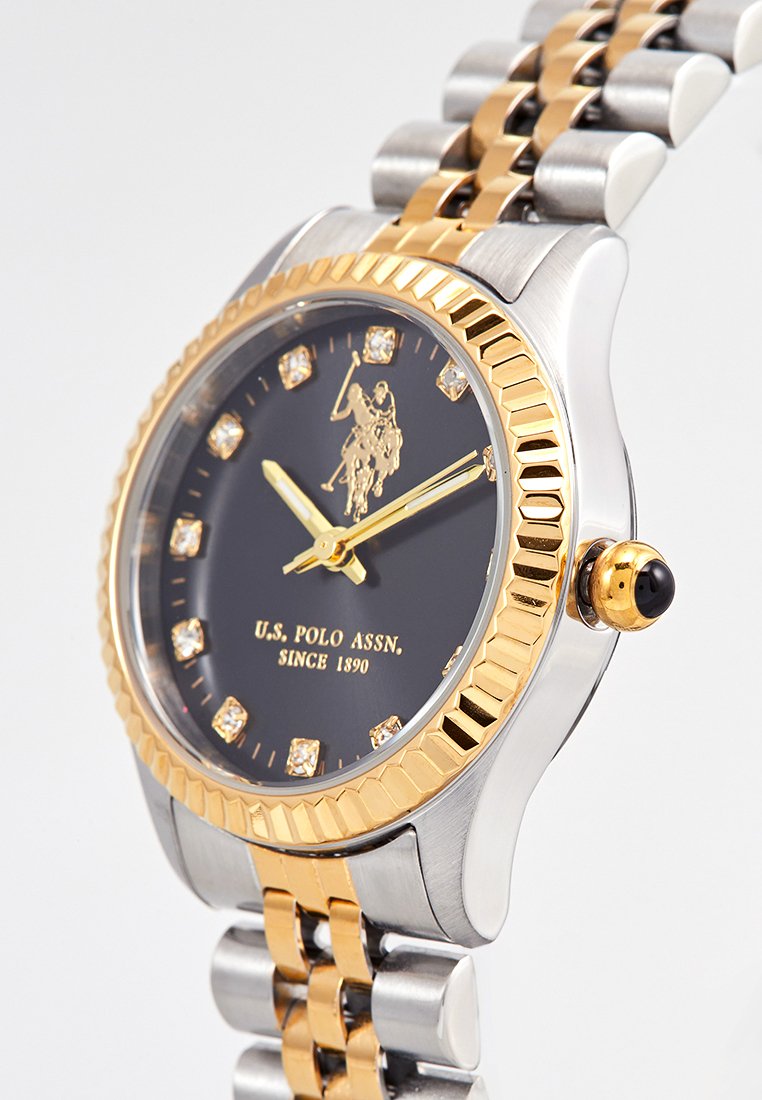 Gold- und silberfarbene Uhr mit schwarzem Zifferblatt, Edelstein-Stundenmarkierungen und einem Logo. Merkmale sind eine strukturierte Lünette und ein farblich abgestimmtes Metallarmband.