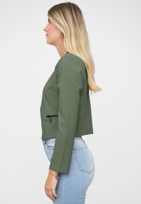 Olijfgroene cropped jas met lange mouwen, een ronde halslijn en een ritszakdetail aan de linkerkant, gedragen met lichtblauwe spijkerbroeken.