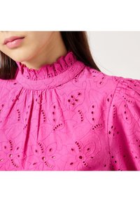 Blusa rosa ricamata con alto colletto a volant, motivo floreale e dettagli in pizzo. Maniche corte a sbuffo e tessuto di cotone testurizzato.
