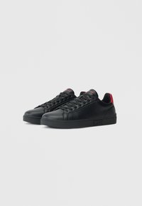 Adidași negri din piele cu o finisare netedă, panou lateral texturat, talpă neagră, accente roșii la călcâi și șireturi negre.