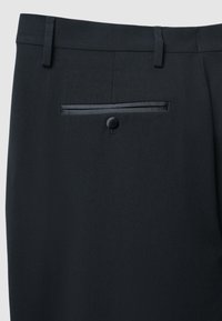 Schwarze, maßgeschneiderte Hose aus glattem Stoff mit einer Gesäßtasche, die mit einem Knopfverschluss und einem satinähnlichen Besatz entlang der Taschenaussparung versehen ist.