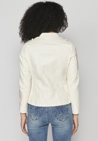 Veste en simili cuir blanche avec des coutures détaillées, épaulettes et accents brodés sur les manches. Associée à un jean bleu.