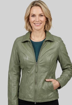 Frau mit kurzen blonden Haaren, die lächelt und eine olivgrüne Lederjacke trägt, die über einem petrolfarbenen Oberteil zugezippt ist, Hand in der Jackentasche.