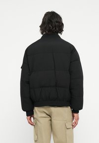 Schwarze gepolsterte Jacke mit hohem Kragen, oversized Passform, gerippten Bündchen und Saum. Der Stoff wirkt glatt mit einem matten Finish.