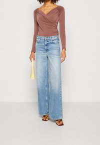 Brauner Langarm-Body mit drapiertem Design, kombiniert mit hellblauen weiter geschnittenen Jeans. Accessoiert mit einer gelben Handtasche und braunen Sandalen.