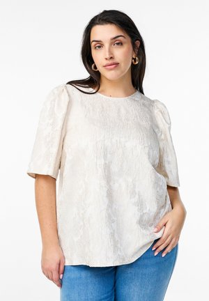 Cream puff-sleeve top with textured fabric, round neckline, and a loose fit. Worn with light blue denim jeans.