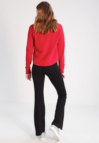 Felpa rossa in misto cotone, abbinata a pantaloni neri a zampa d'elefante. Il modello indossa sneaker argentate. Vista da dietro, capelli lunghi e mossi in evidenza.