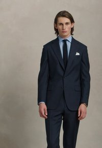 Costume bleu marine au coupe ajustée, associé à une chemise habillée bleu clair et une cravate foncée. Comprend un mouchoir blanc, un tissu lisse et des boutons.