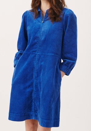 Femme portant une robe en velours côtelé bleu roi, longueur genoux, manches longues, avec une fermeture éclair à l'avant et des poches latérales, debout devant un fond uni.