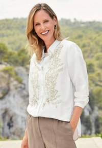 Camicia bianca con ricami di perline dorate sul davanti, con colletto classico e maniche lunghe, abbinata a pantaloni beige.