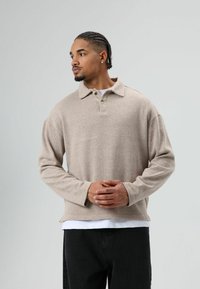 Polo beige à manches longues avec une surface texturée, patte à deux boutons et ourlet incurvé, porté sur une chemise blanche et un pantalon noir.