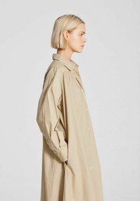 Robe chemise en coton beige avec une coupe décontractée, boutonnière devant, deux poches latérales et manches longues, exhibant une texture lisse et légère.