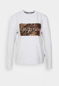 Jasnoszary sweter z bawełny, z metaliczną brązową łatą z cekinów w kształcie prostokąta z napisem "CALVIN KLEIN" w białym kolorze. Standardowy krój.