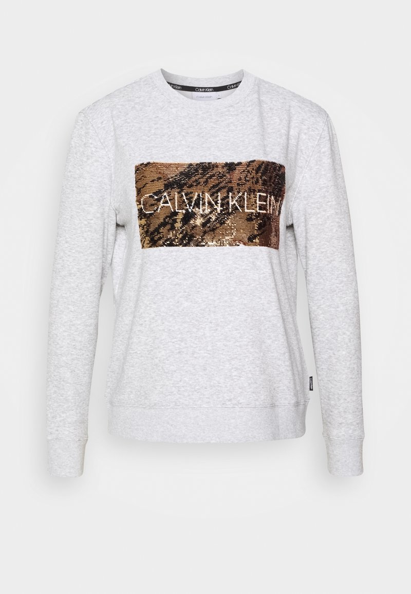 Jasnoszary sweter z bawełny, z metaliczną brązową łatą z cekinów w kształcie prostokąta z napisem "CALVIN KLEIN" w białym kolorze. Standardowy krój.