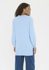 Kvinna med curly hår som bär en långärmad ljusblå tröja och mörkblå jeans, vänd något åt sidan mot en vit bakgrund.